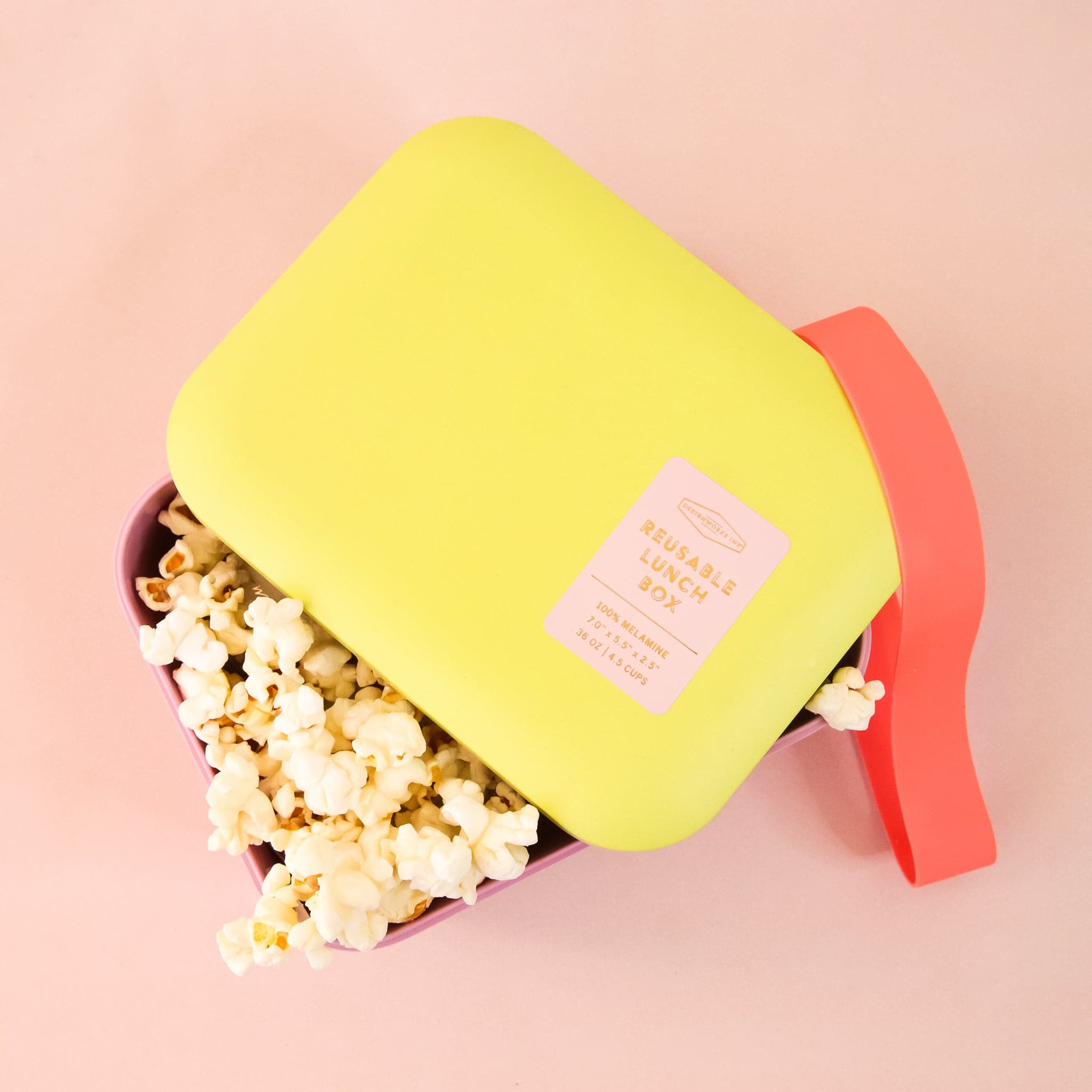 A bright pink and bright yellow lunch box with a red silicone band that goes around to keep closed photographed here with popcorn inside.