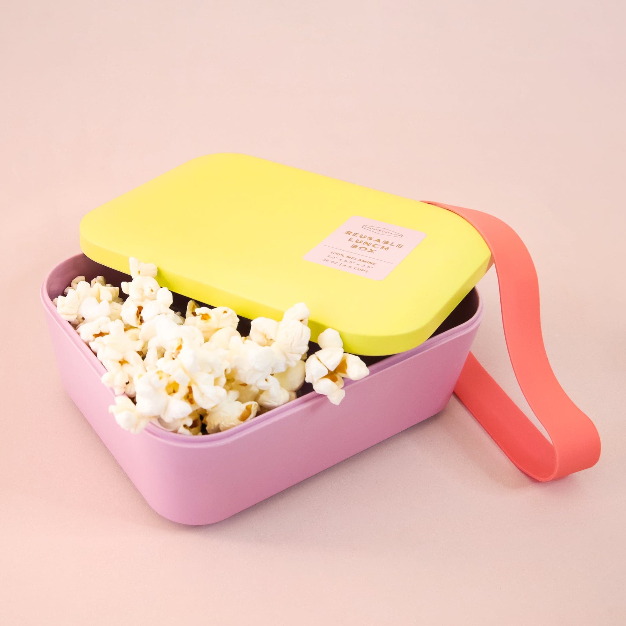 A bright pink and bright yellow lunch box with a red silicone band that goes around to keep closed photographed here with popcorn inside. 