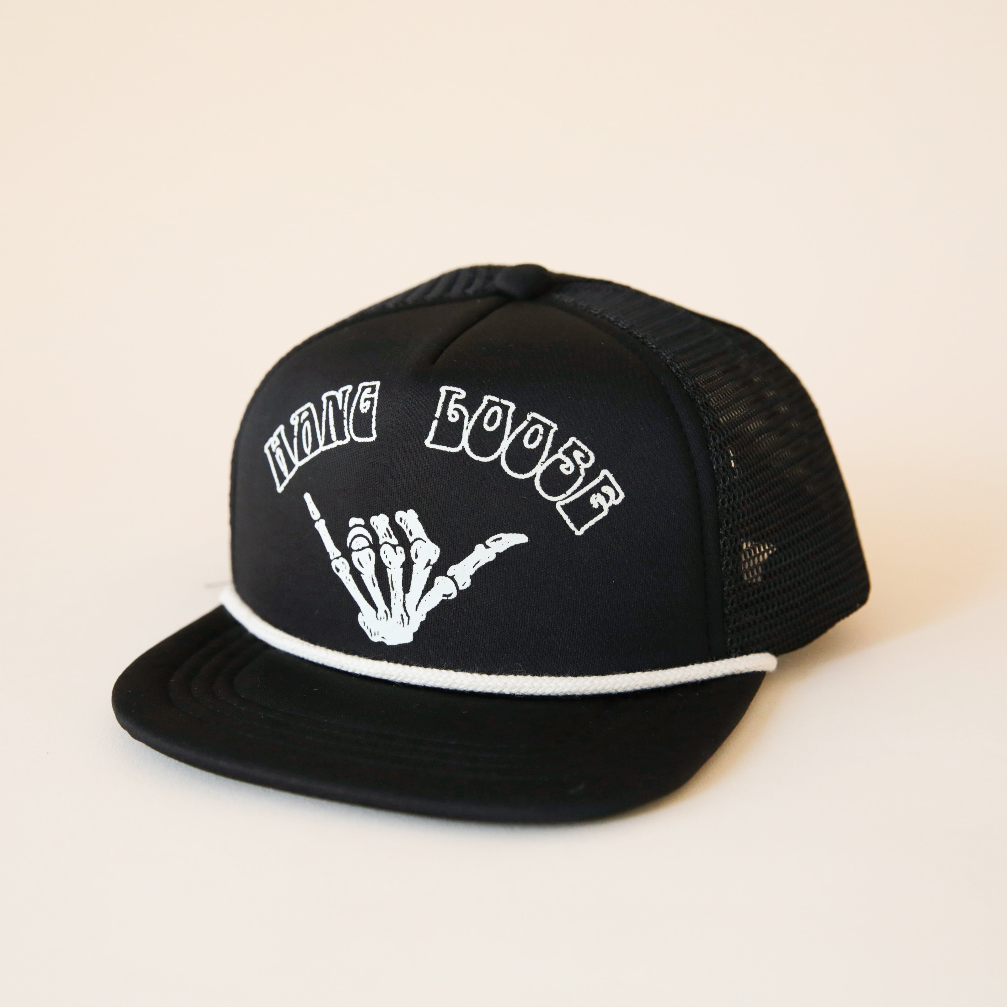 On a cream background is a black foam trucker hat with a white rope detail and white letters arched across the front that read, &quot;Hang Loose&quot; along with a skeleton hand throwing up hang loose. 