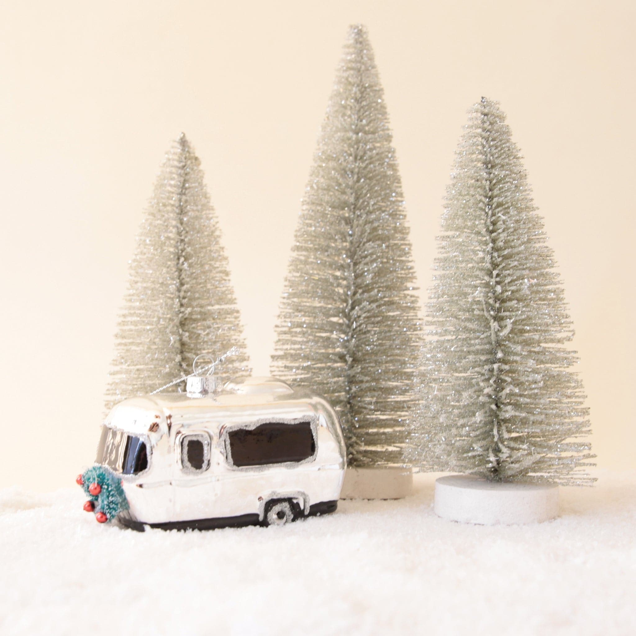 a silver camper ornament with silver bristle trees of varying sizes behind it all sitting on snow with a neutral backgorund