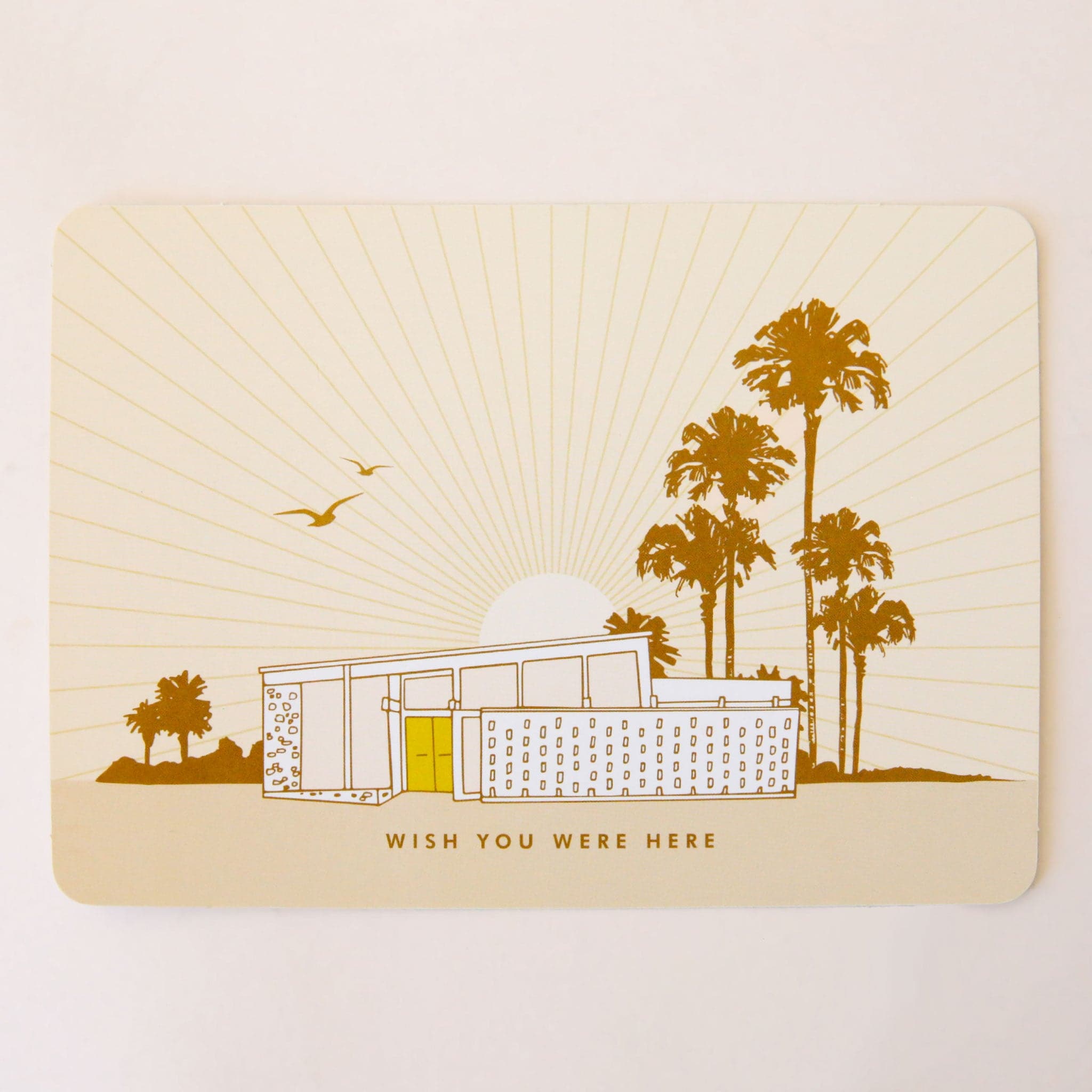 A light pink post card with an illustration of a mid century home along with palm trees and a sun behind the house. The small text at the bottom reads, &quot;Wish you were here&quot;.