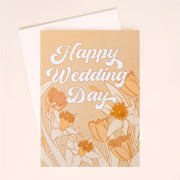 A pink card with white bubble cursive text that reads, "Happy Wedding Day" along with tulip and daffodil illustrations along the bottom. The card also comes with a white envelope.