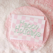 Pink and white checkered greeting card that reads 'Happy Holidays' in white font with a thin green shadow. The card is accompanied by a solid white envelope.