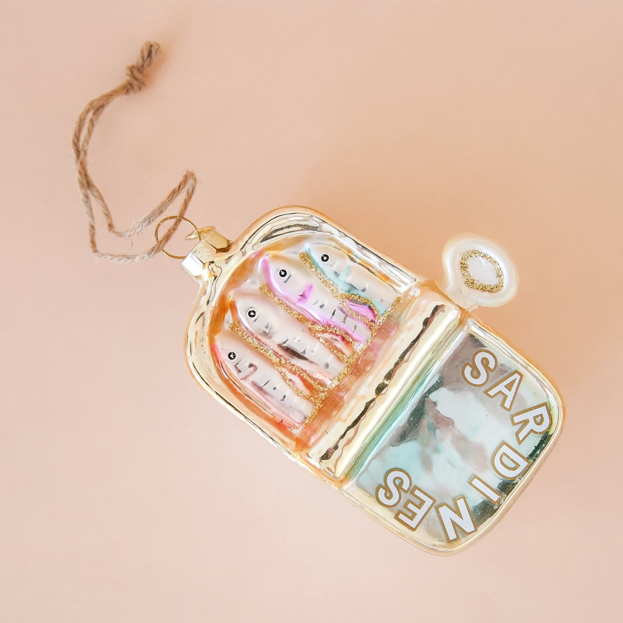 In front of a peachy background is a rounded rectangular glass ornament. The ornament is gold metallic. The bottom half is a silvery blue with white text that is outlined in gold. The text reads ‘sardines.’ The bottom half is four heads of sardines. On the right side of the ornament is a white and gold handle. On the top end of the ornament is a gold hook with brown twine. 