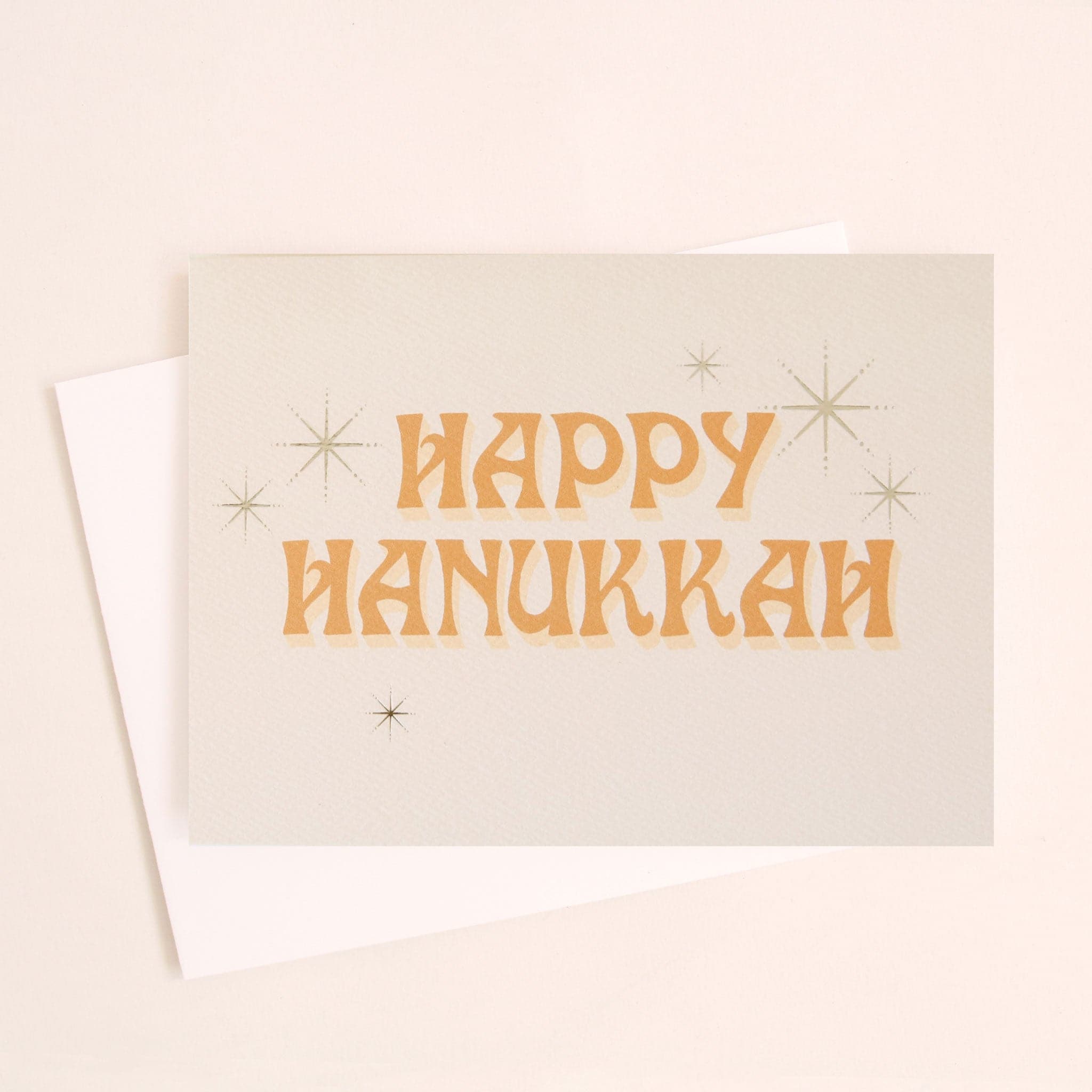 Ivory card that reads &#39;Happy Hanukkah&#39; in retro orange lettering. The text is accented with gold foil stars twinkling around.