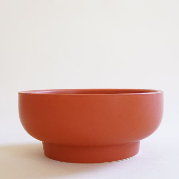 In front of a white background is a rust colored pedestal bowl. The top is a wide and short bowl with a short pedestal bottom. 