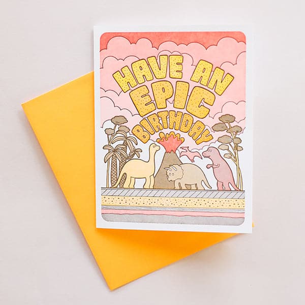 On a white background is a yellow and orange card with dinosaurs and a volcano graphic along with text arched over the illustration that says, "Have An Epic Birthday" along with a coordinating yellow envelope. 