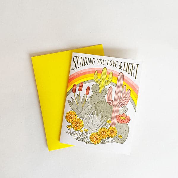 On top of a yellow envelope is a white card. At the top is brown text that reads ‘sending you love & light.’ Below is a rainbow that is pink, red, orange, yellow and green. Below is a variety of cacti and succulents. 