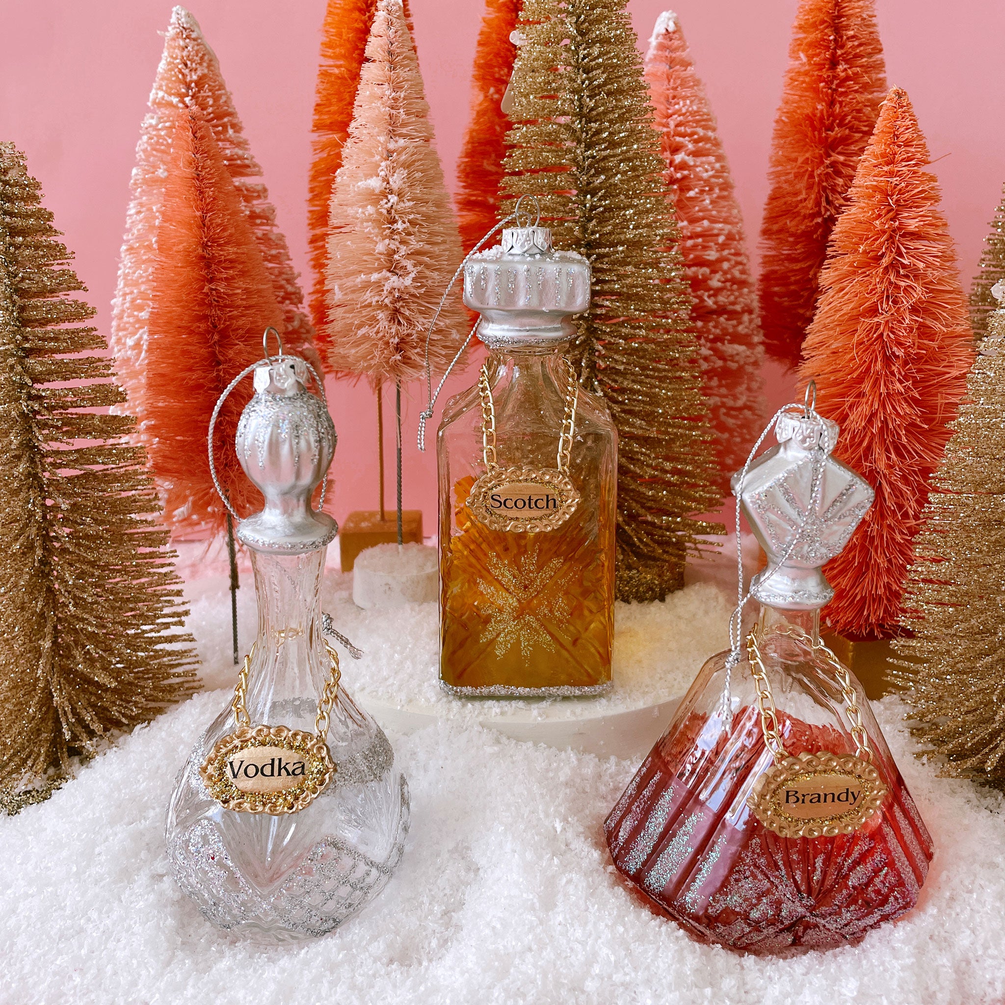 Three glass decanter ornaments with a gold label that reads, "Brandy", "Scotch", and "Vodka" along with a pinkish base. The stoppers to the decanters are silver and detailed with glitter.