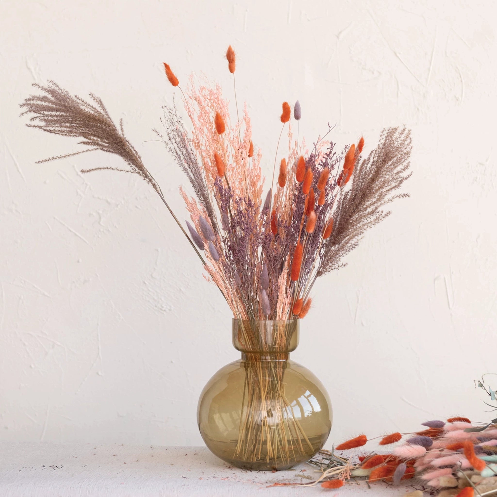 A bundle of dried bunny tail florals in a rich coral shade with long stems photographed here in an arrangement in a green vase.