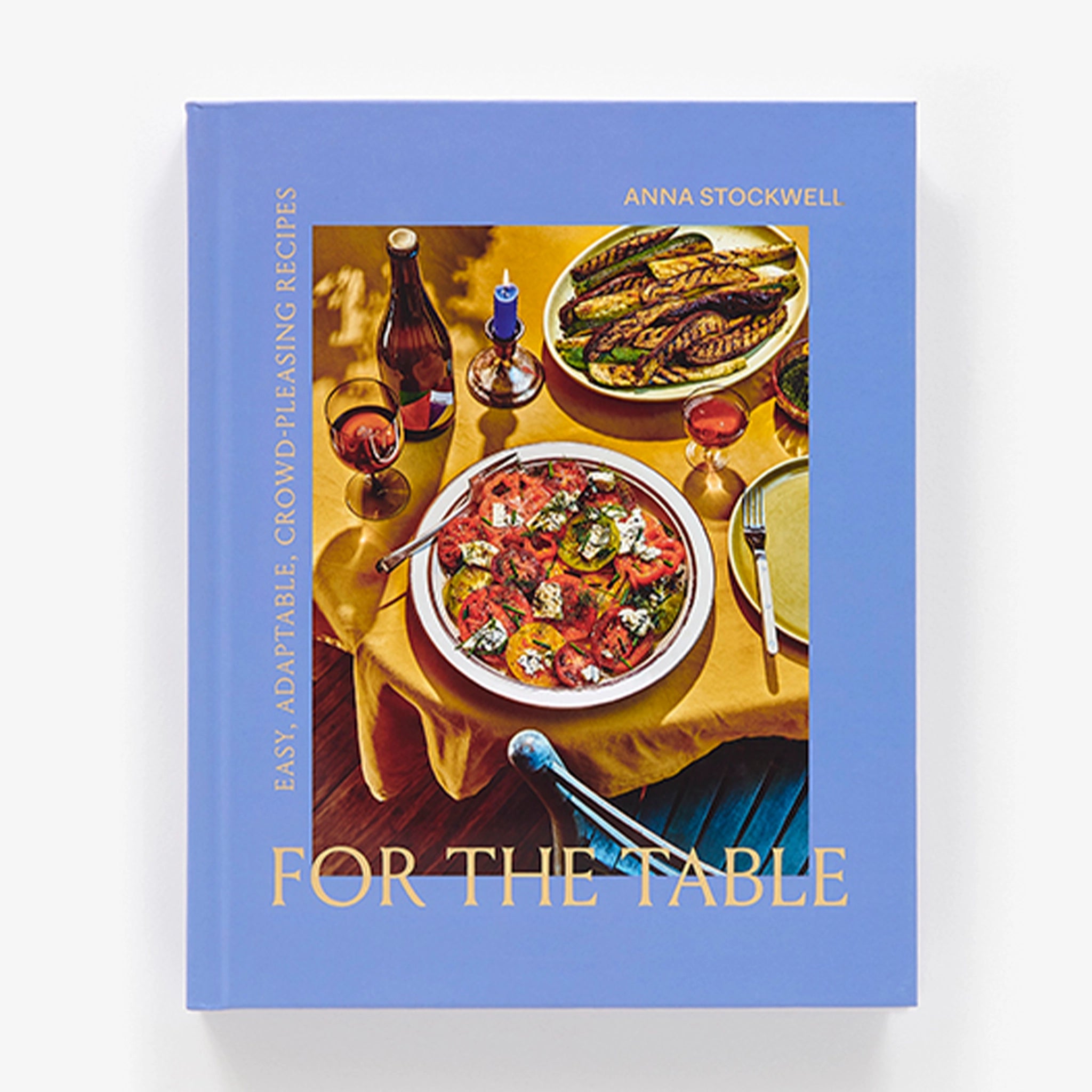 A indigo blue book cover with a photograph of a beautiful table scape filled with different dishes of food and wine as well as the title in pale yellow that reads, "For The Table Easy, Adaptable, Crow-Pleasing Recipes". 