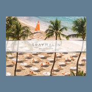 A boxed puzzle with a seaside aerial photograph from Gray’s best-selling collection, À la Plage, featuring a beach with umbrellas, sun chairs and palm trees.