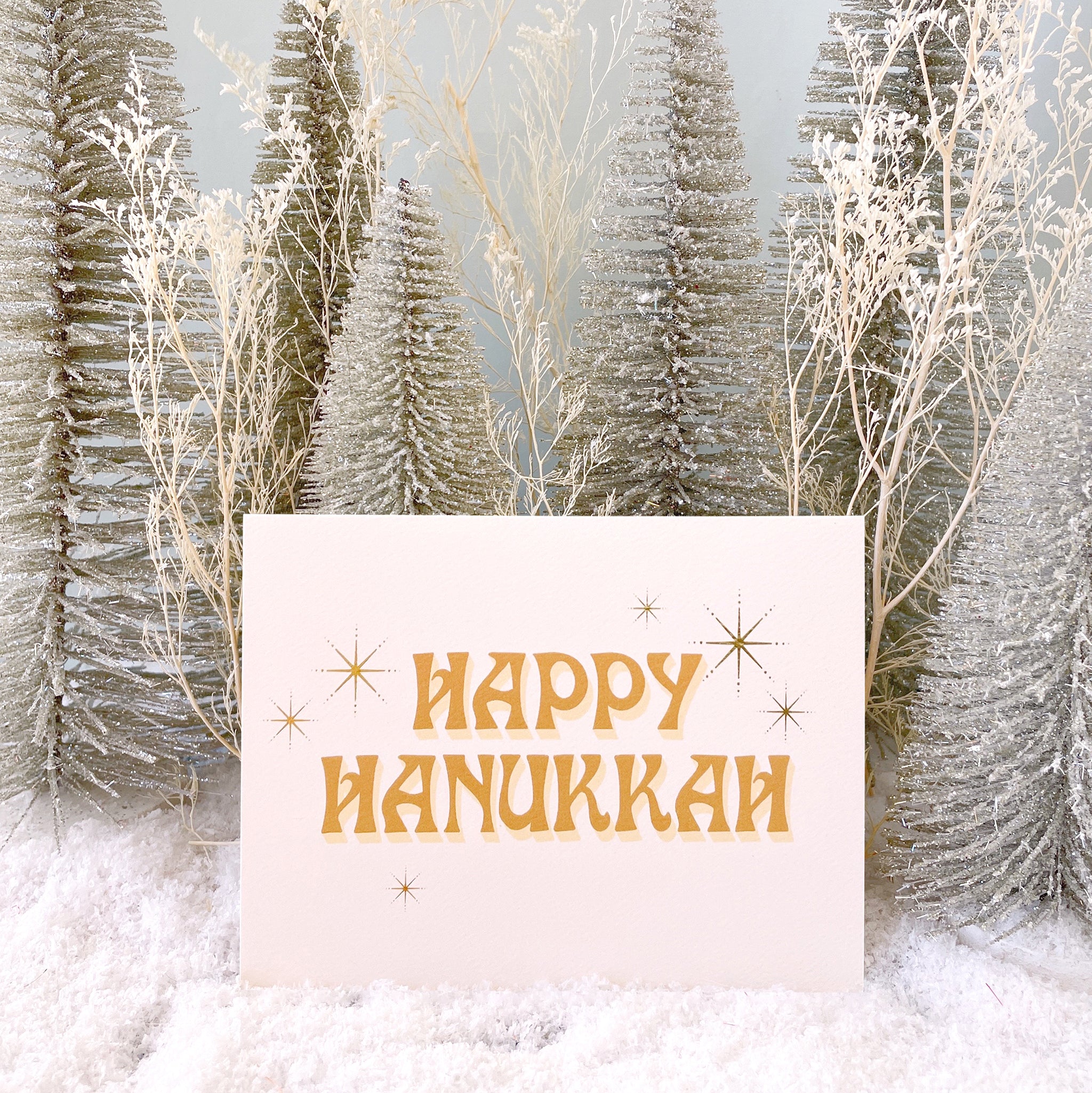 Ivory card that reads &#39;Happy Hanukkah&#39; in retro orange lettering. The text is accented with gold foil stars twinkling around.