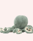 In front of a white background is a light blue stuffed octopus. On his head are two black eyes and a small black mouth. The bottom of his tentacles is cream colored. 