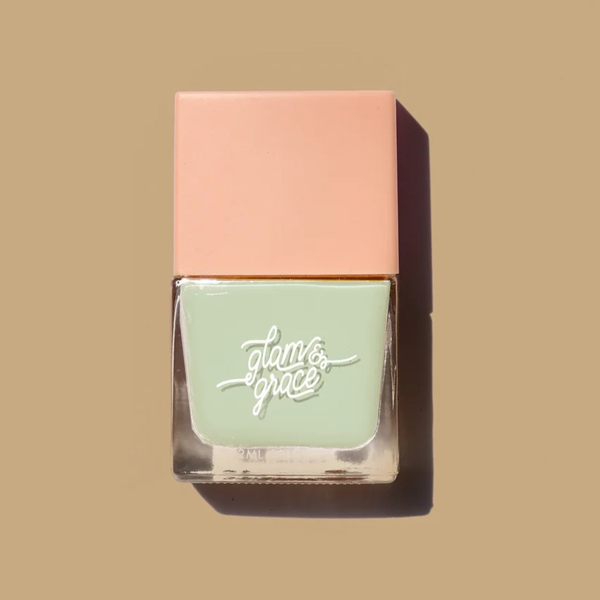 A mint green nail polish in a square glass jar with a salmon pink lid as well as white cursive text that reads, "glam & grace".