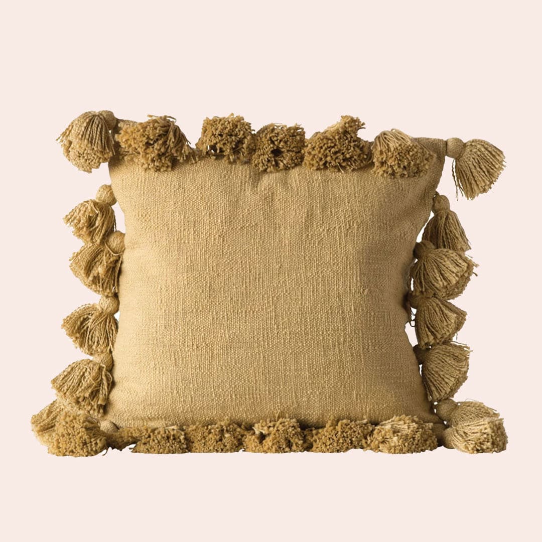 On a cream background, is a mustard yellow pillow with tassels from all alone the edges of the square pillow.