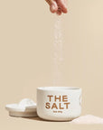 A white ceramic jar with a hand dropping medium grind salt into the jar along with a half moon lid off to the side.