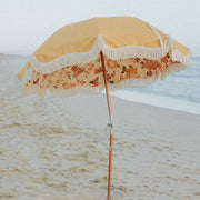 This is a picture of a sandy beach. In the middle of the sand is an umbrella. The umbrella has a wood post. The top of the umbrella is muted yellow and the inside is white with a dark orange and brown paisley design. The bottom of the umbrella is bordered with cream tassels. 