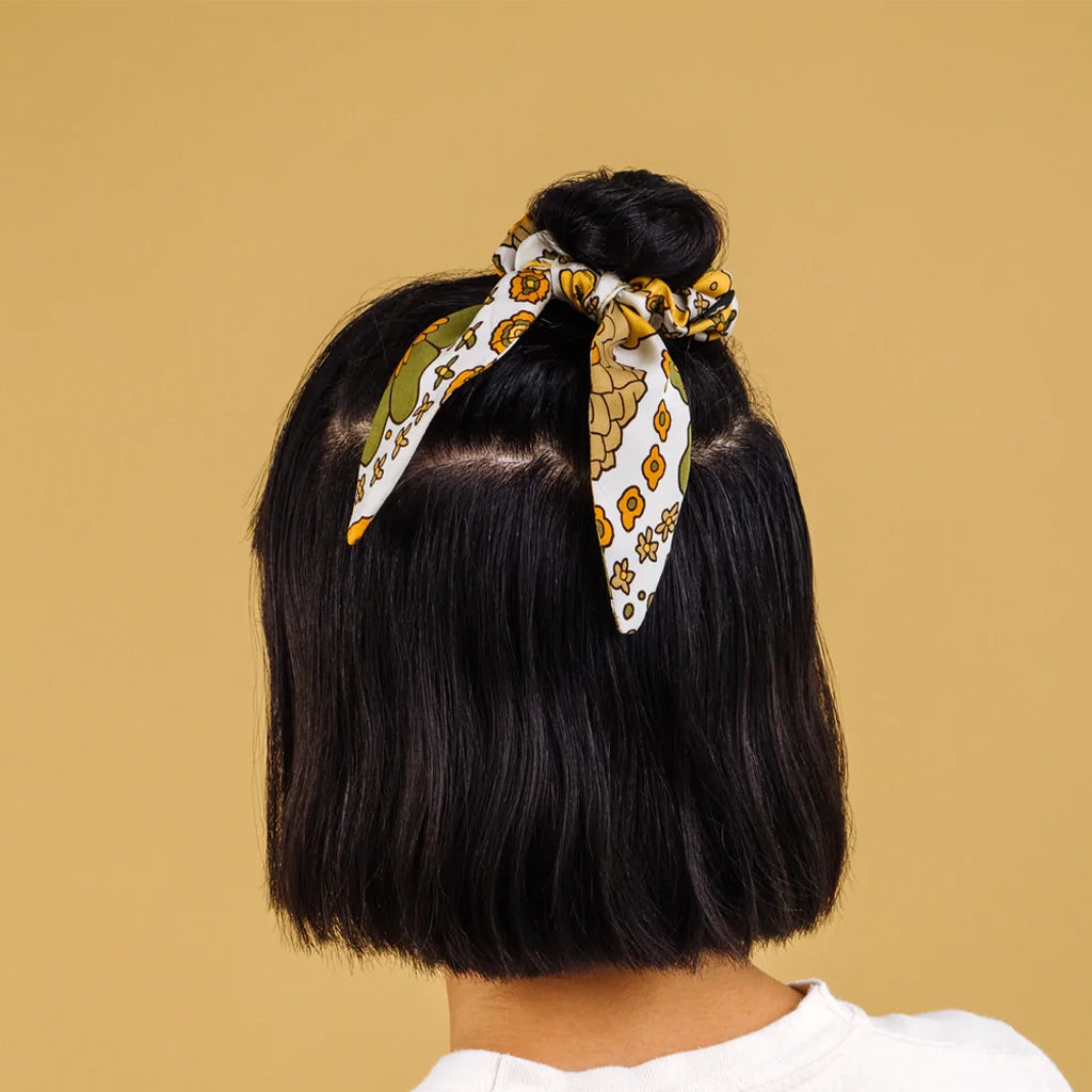 A satin scrunchie hair tie that has a yellow, olive green and white floral pattern on it photographed here being worn on a model with a half up half down hairstyle.