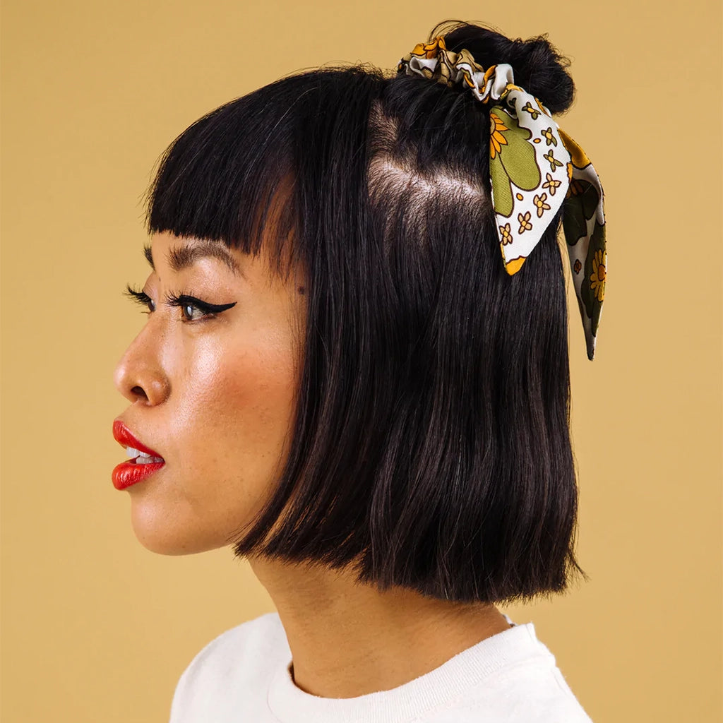 A satin scrunchie hair tie that has a yellow, olive green and white floral pattern on it photographed here being worn on a model with a half up half down hairstyle.