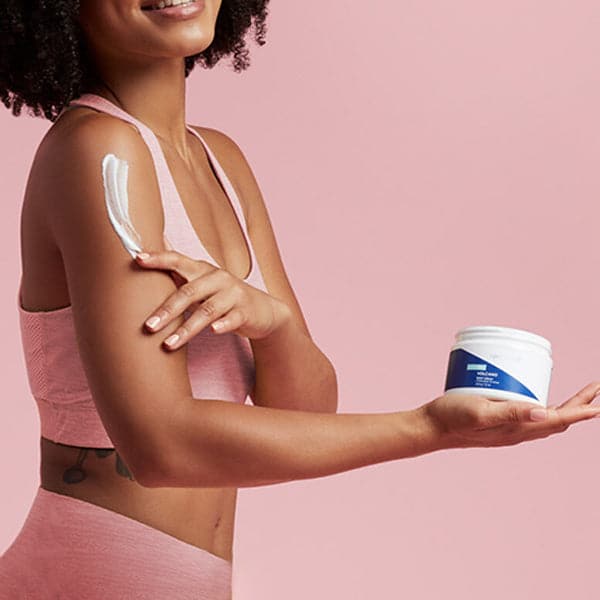 Against a rose pink background stands a woman with the side of her body in view. She is wearing a matching pink sports bra and workout bottoms. In one hand she is holding a plastic white jar. On the body of the jar there is a sticker wrap that is split diagonally by color. The top half is white and the bottom half is cobalt blue. The lid is not on the jar. The model is using her other hand to rub white lotion on her arm that is holding the jar. 