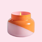 A squatty, round glass jar sits in front of a white background. The jar is split diagonally with the top half being a bright orange color and the bottom half being a soft pink color. The top of the jar is a lot narrower than the rest of the jar. The inside of the jar is the same shade of pink as the outside of the jar. There is a small flame inside the candle. 